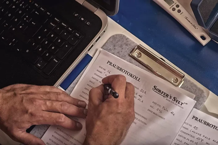 Ein Brauer dokumentiert sorgfältig den Brauprozess von Sorfer's Sylt auf einem Protokollbogen neben einem Laptop für die handwerkliche Qualität.