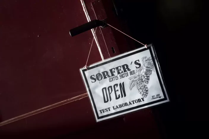 Ein Schild mit der Aufschrift Sorfer's Echtes Sylter Bier Open Test Laboratory hängt an einer dunklen Tür und lädt herzlich zum Besuch ein.