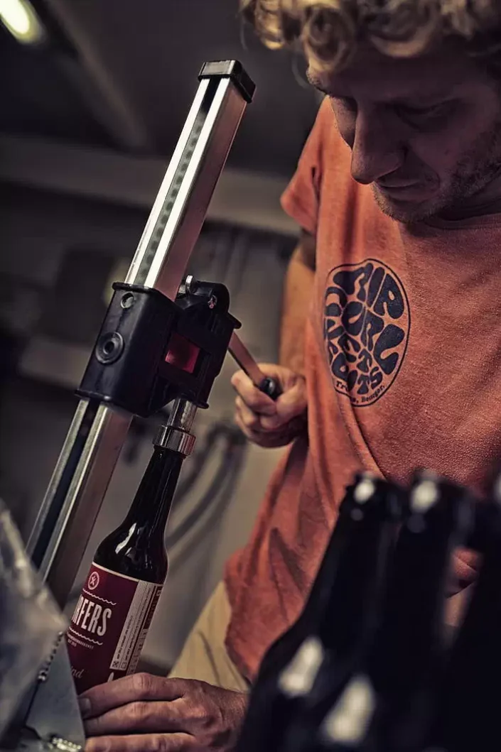 Ein konzentrierter Mann in einem orangefarbenen T-Shirt verschließt sorgfältig eine Bierflasche mit einer manuellen Abfüllmaschine.
