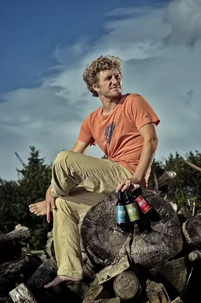 Ein entspannter Mann mit lockigem Haar sitzt barfuß auf einem Holzstapel und hält drei Flaschen Surfers Bier in der Hand.