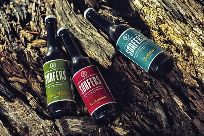 Drei Flaschen Sørfers Bier mit bunten Etiketten liegen auf einem rustikalen, rauen Baumstamm in der Natur von Sylt.