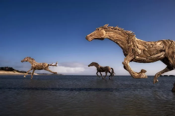 Drei lebensgroße Treibholzskulpturen von galoppierenden Pferden im flachen Wasser vor der Küste einer Nordseeinsel bei strahlendem Sonnenschein.