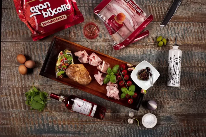 Eine rustikale Holzplatte mit italienischen Spezialitäten wie Gran Biscotto Schinken, frischem Brot, Oliven und Wein auf einem Holztisch.