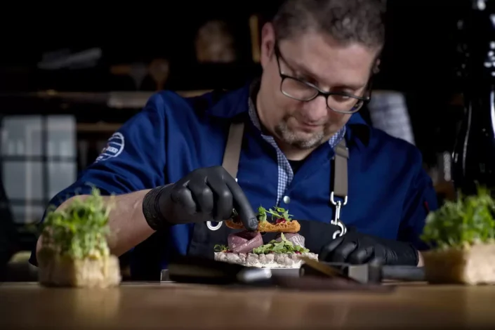 Ein konzentrierter Koch mit Brille und schwarzen Handschuhen richtet kunstvoll ein Gourmet-Gericht an, ideal für kulinarische Portraits.