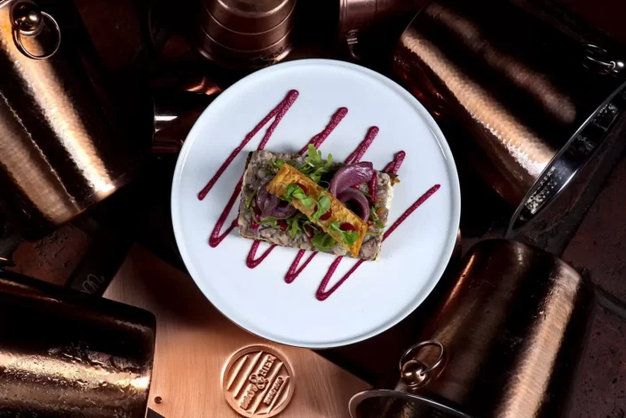 Ein kunstvoll angerichtetes Gericht auf einem weißen Teller, umgeben von glänzenden Kupferbechern, serviert als Teil der Foodfotografie.