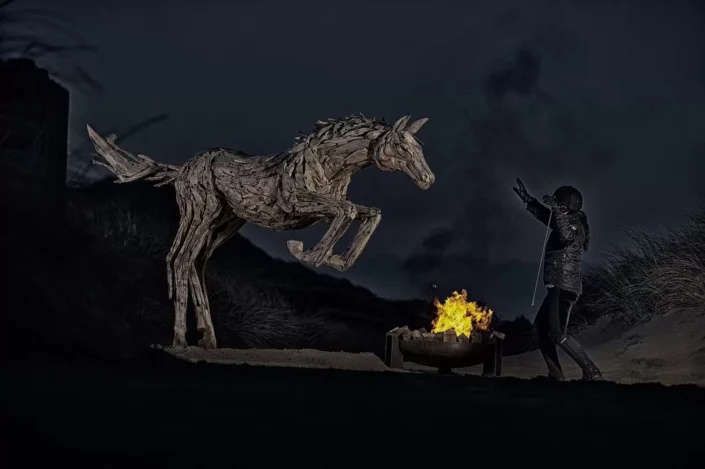 Eine Reiterin interagiert bei Nacht am Strand mit einer kunstvollen Treibholz-Skulptur eines springenden Pferdes neben einem Lagerfeuer.