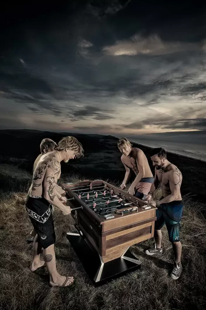 Vier Männer spielen bei dramatischem Abendhimmel an einem Kicker auf einer Düne, ein stimmungsvolles Bild für kreative Lifestyle Fotografie.