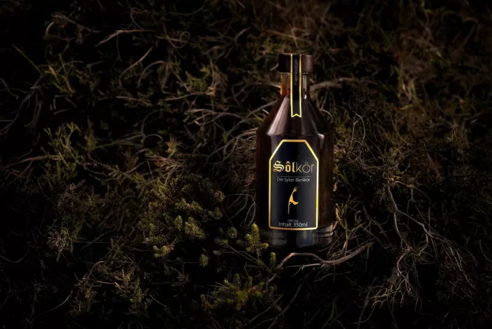 Eine Flasche Sölkör Bierlikör steht stimmungsvoll in dunklem Heidekraut, präsentiert in einer professionellen Produktfotografie.