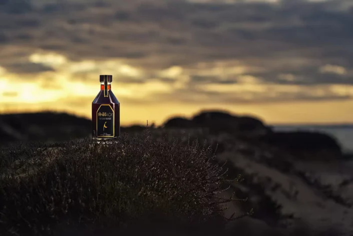 Eine Flasche Sölkör Likör steht stimmungsvoll in der Heide auf Sylt vor einem goldenen Sonnenuntergang am Meer für ästhetische Produktfotografie.