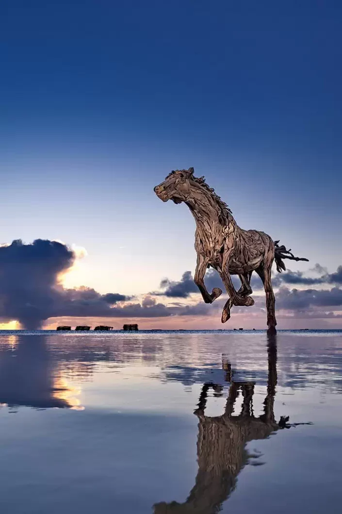 Eine kunstvolle Treibholzskulptur eines galoppierenden Pferdes im flachen Wasser vor einem stimmungsvollen Sonnenuntergang an der Nordseeküste.