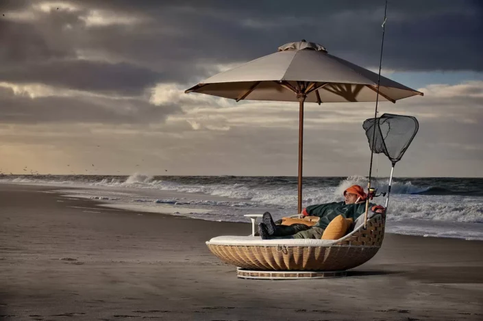 Ein entspannter Angler ruht sich in einem Strandkorb am Meer aus, während im Hintergrund die Wellen unter einem dramatischen Abendhimmel brechen.
