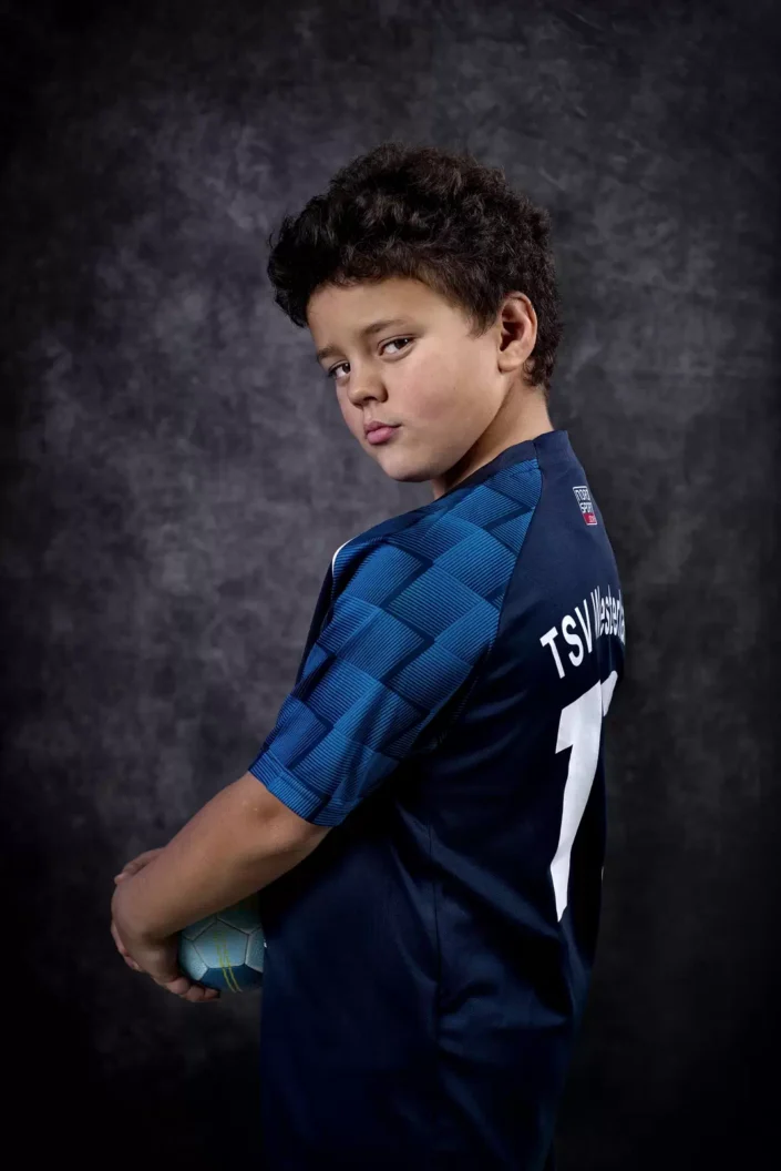 Ein Junge mit dunklen Locken trägt ein blaues Trikot vom TSV Westerland und hält einen Handball vor einem grauen Hintergrund auf Sylt.