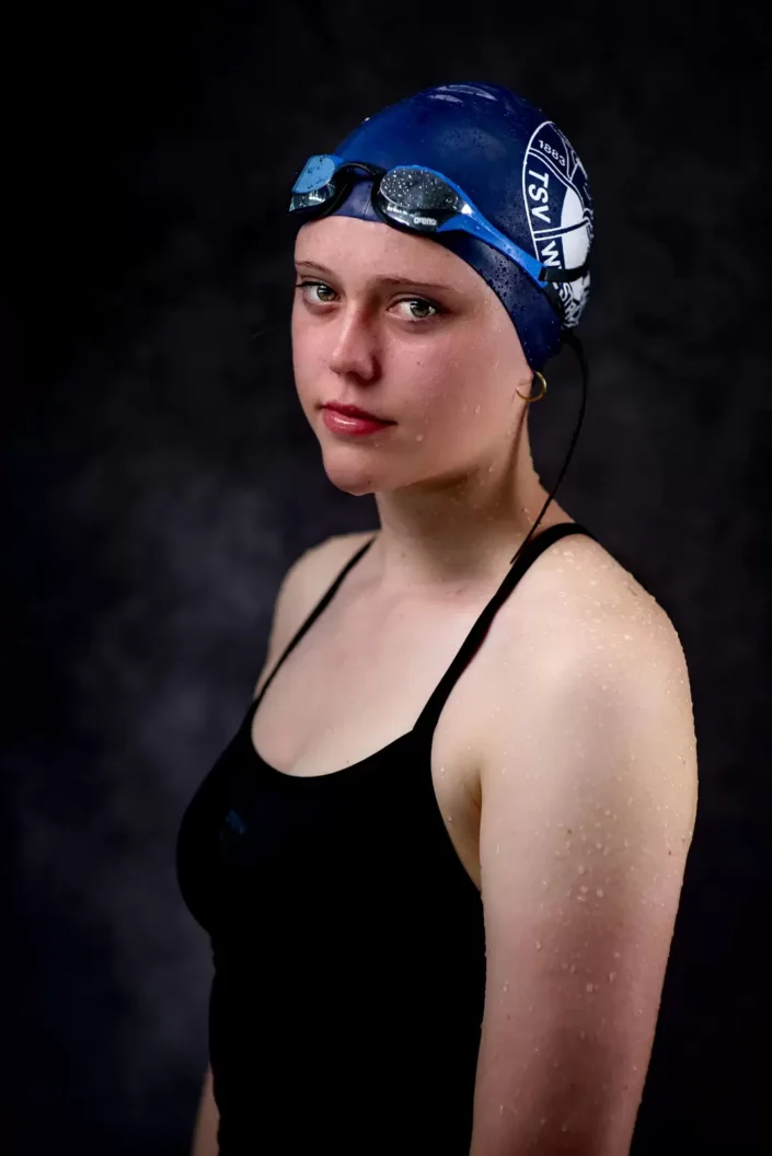 Eine junge Schwimmerin mit blauer Badekappe und Schwimmbrille blickt ernst in die Kamera, ihre Haut ist mit Wassertropfen benetzt.