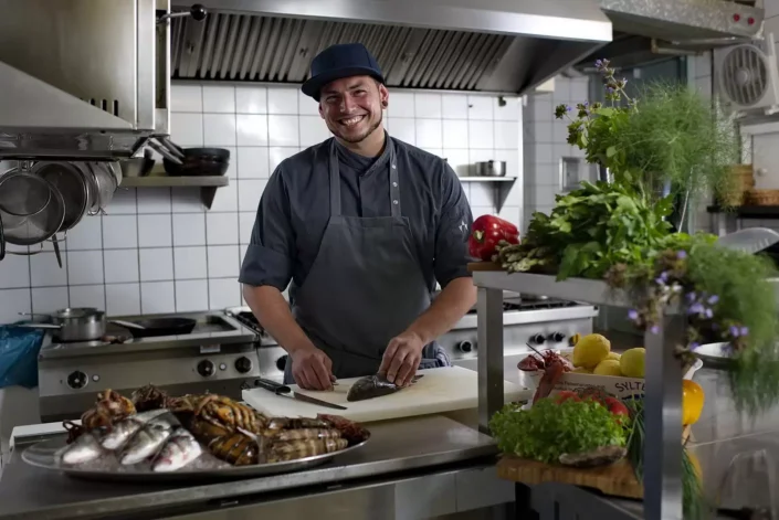 Ein lächelnder Koch in Arbeitskleidung bereitet in einer professionellen Küche auf Sylt frischen Fisch für ein Portrait zu.