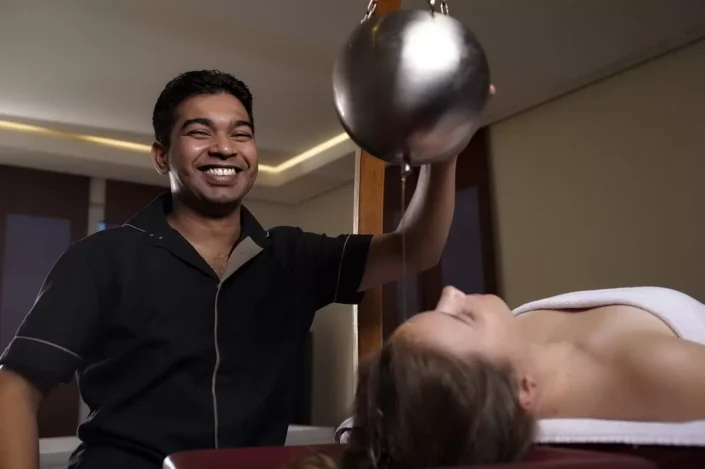 Ein lächelnder Masseur führt eine entspannende Ayurveda-Stirnguss-Behandlung bei einer Kundin in einem Wellness-Studio durch.