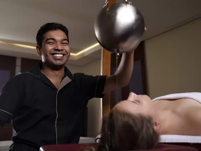 Ein lächelnder Masseur führt eine entspannende Ayurveda-Stirnguss-Behandlung bei einer Kundin in einem Wellness-Studio durch.