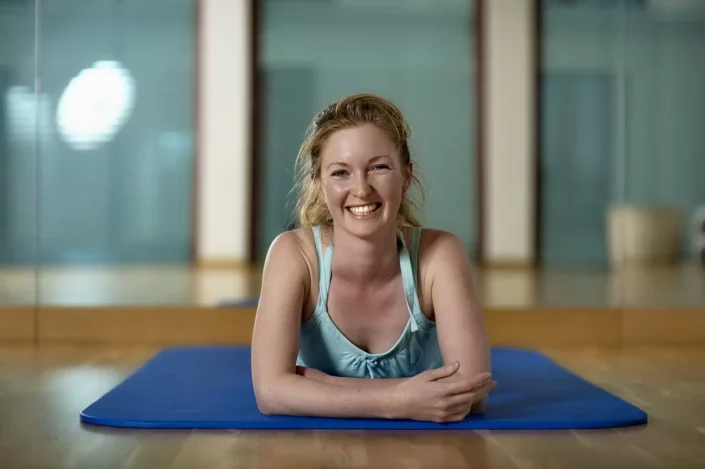 Eine fröhliche Frau in Sportkleidung liegt auf einer blauen Yogamatte in einem hellen Studio für professionelle Portraits und Lifestyle-Fotos.