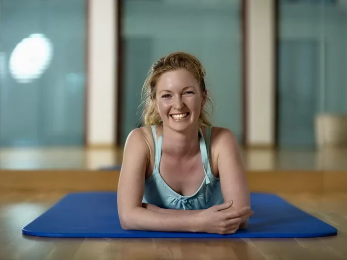 Eine fröhliche Frau in Sportkleidung liegt auf einer blauen Yogamatte in einem hellen Studio für professionelle Portraits und Lifestyle-Fotos.