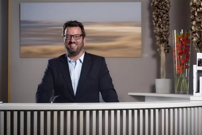 Ein freundlicher Fotograf mit Brille und Sakko lächelt hinter einem Empfangstresen für professionelle Portraits auf Sylt.