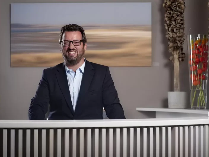 Ein freundlicher Fotograf mit Brille und Sakko lächelt hinter einem Empfangstresen für professionelle Portraits auf Sylt.