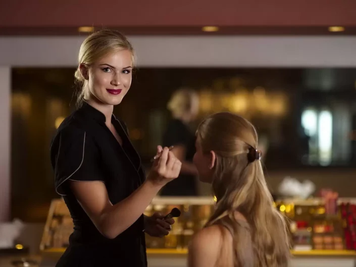Eine Visagistin trägt bei der professionellen Hochzeitsfotografie auf Sylt Make-up auf, während die Braut geduldig im Hintergrund sitzt.