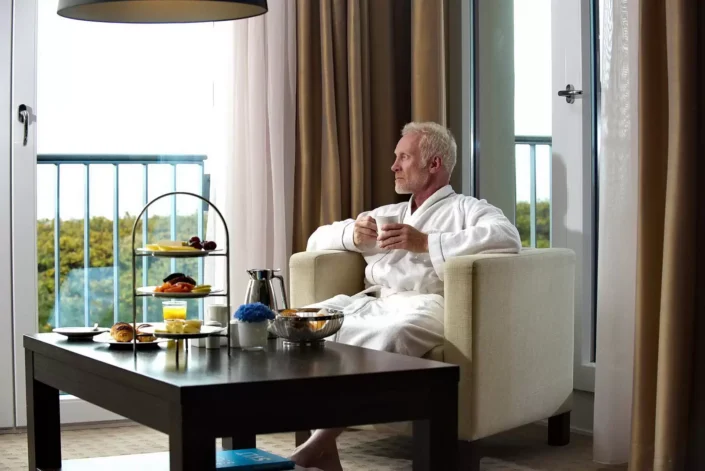 Ein entspannter Mann im Bademantel sitzt mit einer Tasse Kaffee in einem Hotelzimmer auf Sylt und genießt den Ausblick.