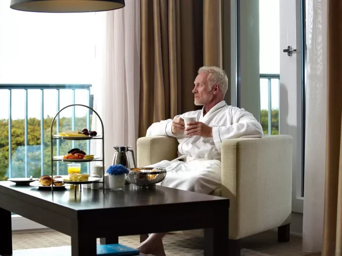 Ein entspannter Mann im Bademantel sitzt mit einer Tasse Kaffee in einem Hotelzimmer auf Sylt und genießt den Ausblick.