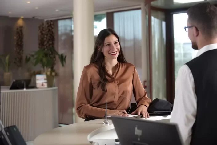 Eine freundliche Frau lächelt an einer Hotelrezeption, während sie mit einem Mitarbeiter spricht. Professionelle Business-Portraits.