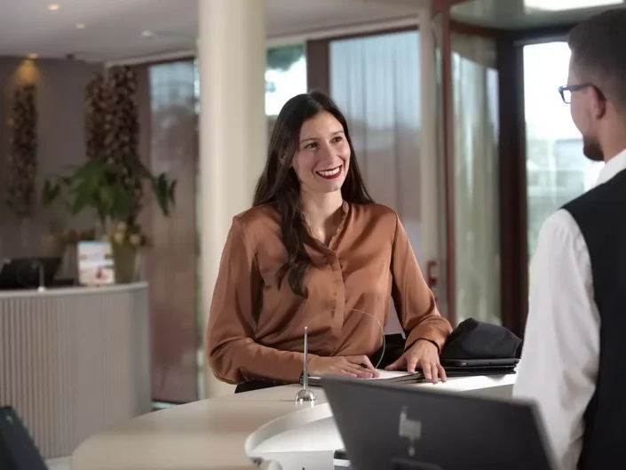Eine freundliche Frau lächelt an einer Hotelrezeption, während sie mit einem Mitarbeiter spricht. Professionelle Business-Portraits.