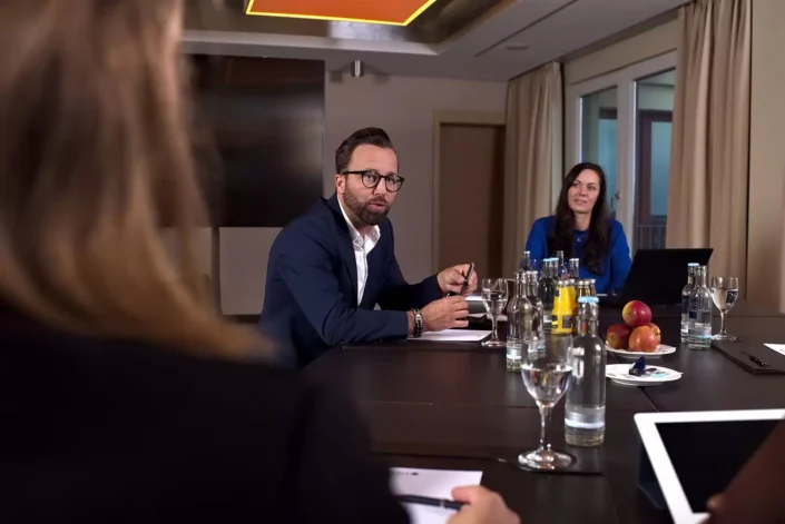 Ein Geschäftsmann mit Brille spricht in einer Konferenzrunde, während eine Kollegin zuhört. Professionelle Business-Fotografie in einem Tagungsraum.