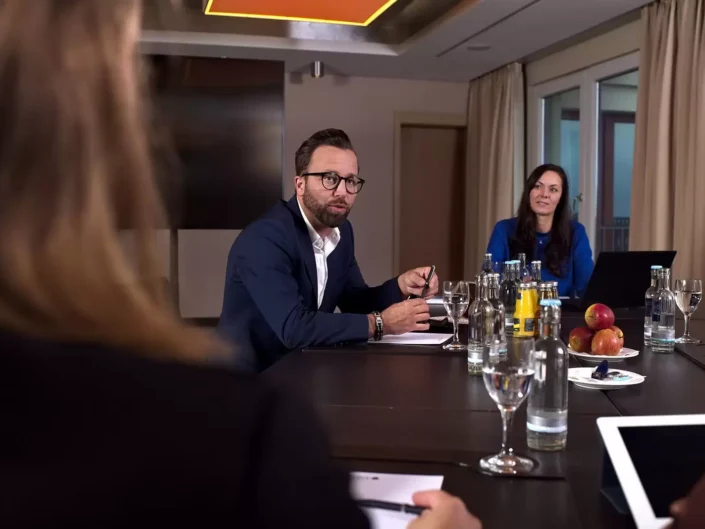 Ein Geschäftsmann mit Brille spricht in einer Konferenzrunde, während eine Kollegin zuhört. Professionelle Business-Fotografie in einem Tagungsraum.