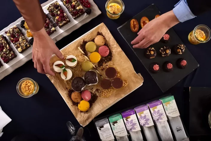 Draufsicht auf ein Business-Catering mit bunten Macarons, Gebäck und Tee auf Sylt, präsentiert von Händen bei der Auswahl der Speisen.