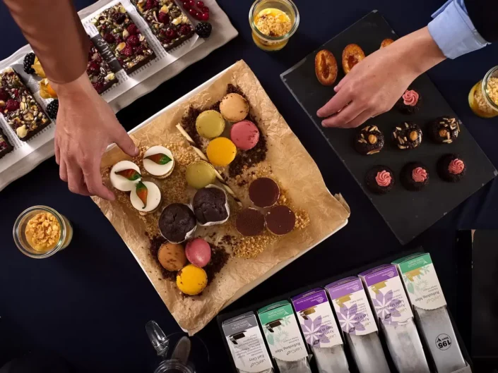 Draufsicht auf ein Business-Catering mit bunten Macarons, Gebäck und Tee auf Sylt, präsentiert von Händen bei der Auswahl der Speisen.