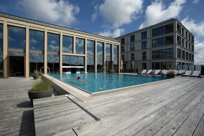 Ein luxuriöser Außenpool vor einem modernen Hotelgebäude auf Sylt, in dem eine Person schwimmt, umgeben von einer hölzernen Terrasse.