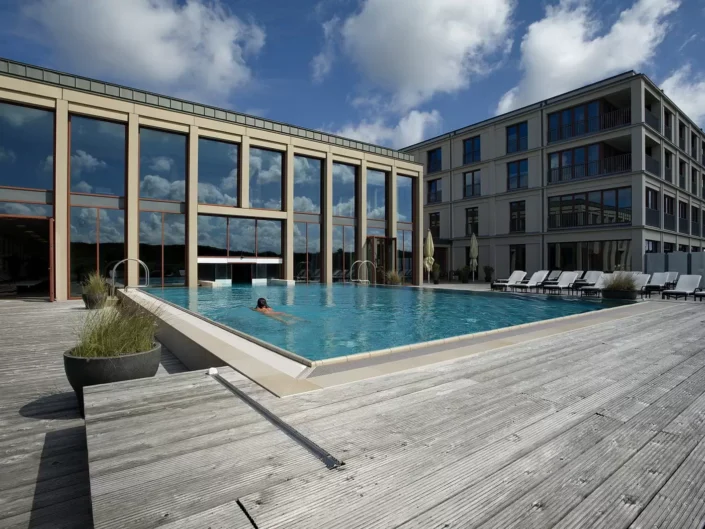 Ein luxuriöser Außenpool vor einem modernen Hotelgebäude auf Sylt, in dem eine Person schwimmt, umgeben von einer hölzernen Terrasse.