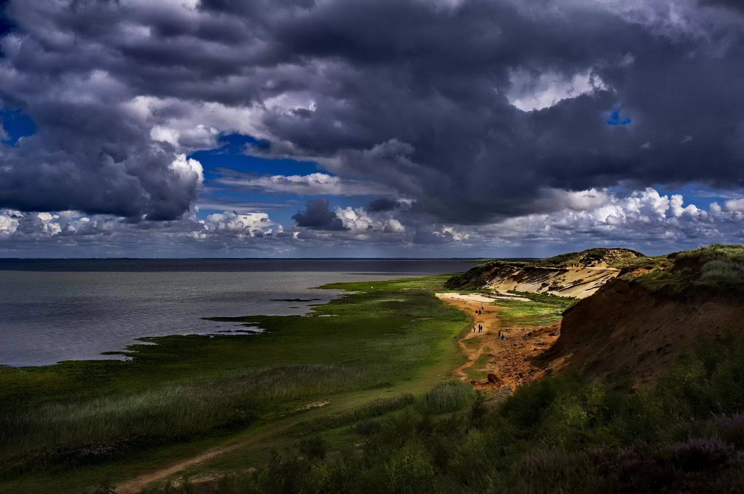 Die dramatische Steilküste von Morsum Kliff auf Sylt mit grünen Wiesen, sandigen Klippen und einem tiefblauen, wolkenverhangenen Himmel über dem Wattenmeer.