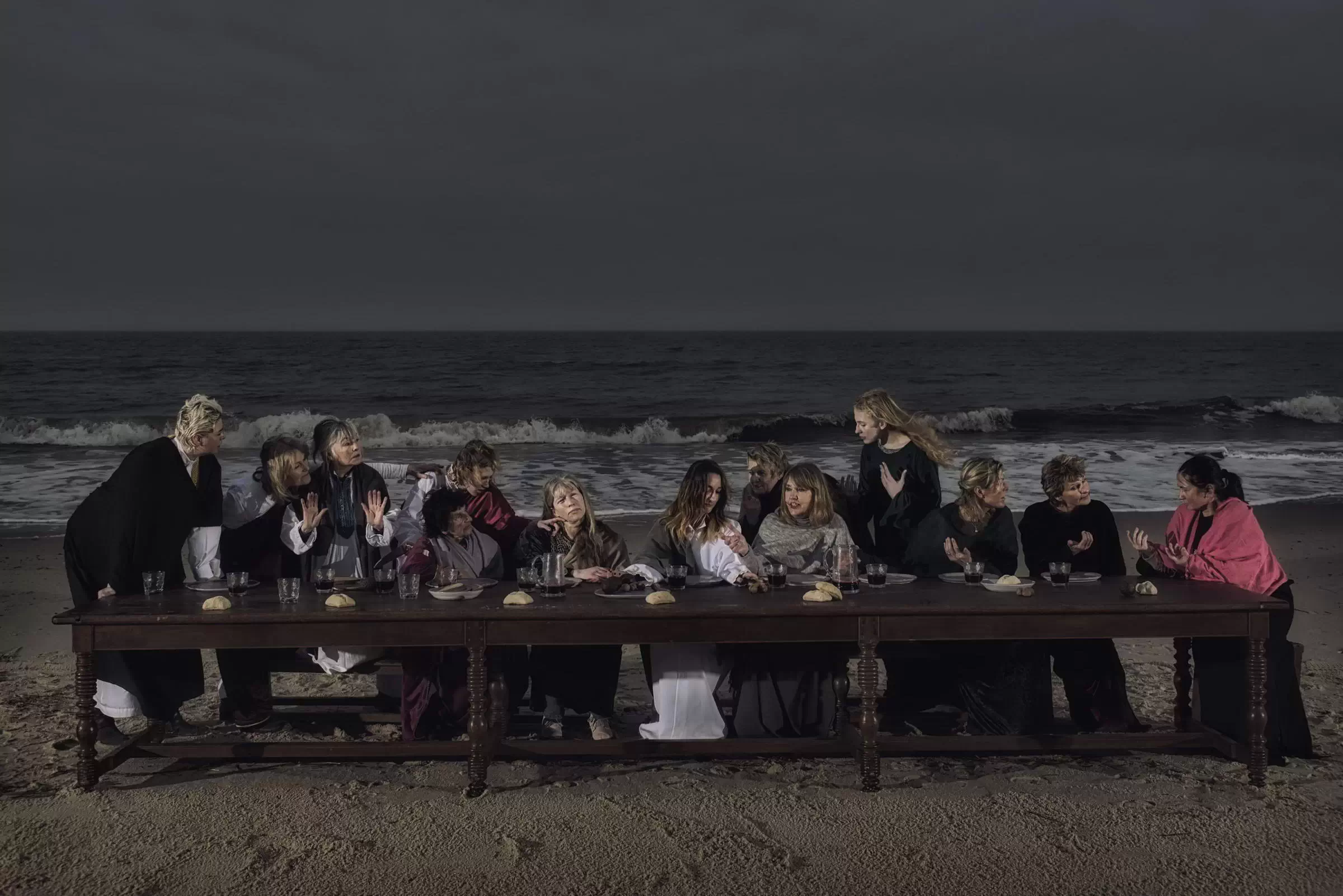 Eine Gruppe von Frauen sitzt an einem langen Holztisch am Strand von Sylt und inszeniert das Abendmahl vor der Kulisse des dunklen Nordseemeeres.