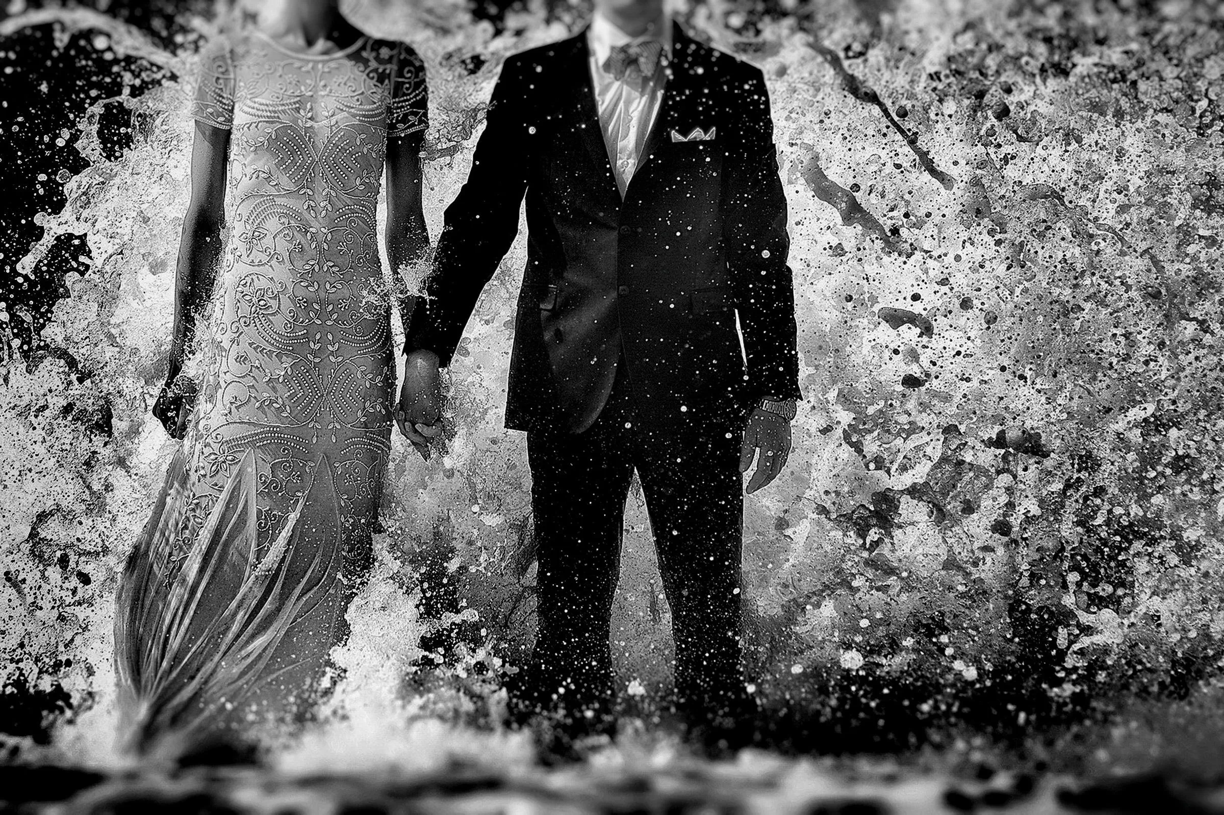 Ein Brautpaar in eleganter Hochzeitskleidung steht händehaltend vor einer imposanten, spritzenden Meereswelle an einem Sylter Strand.