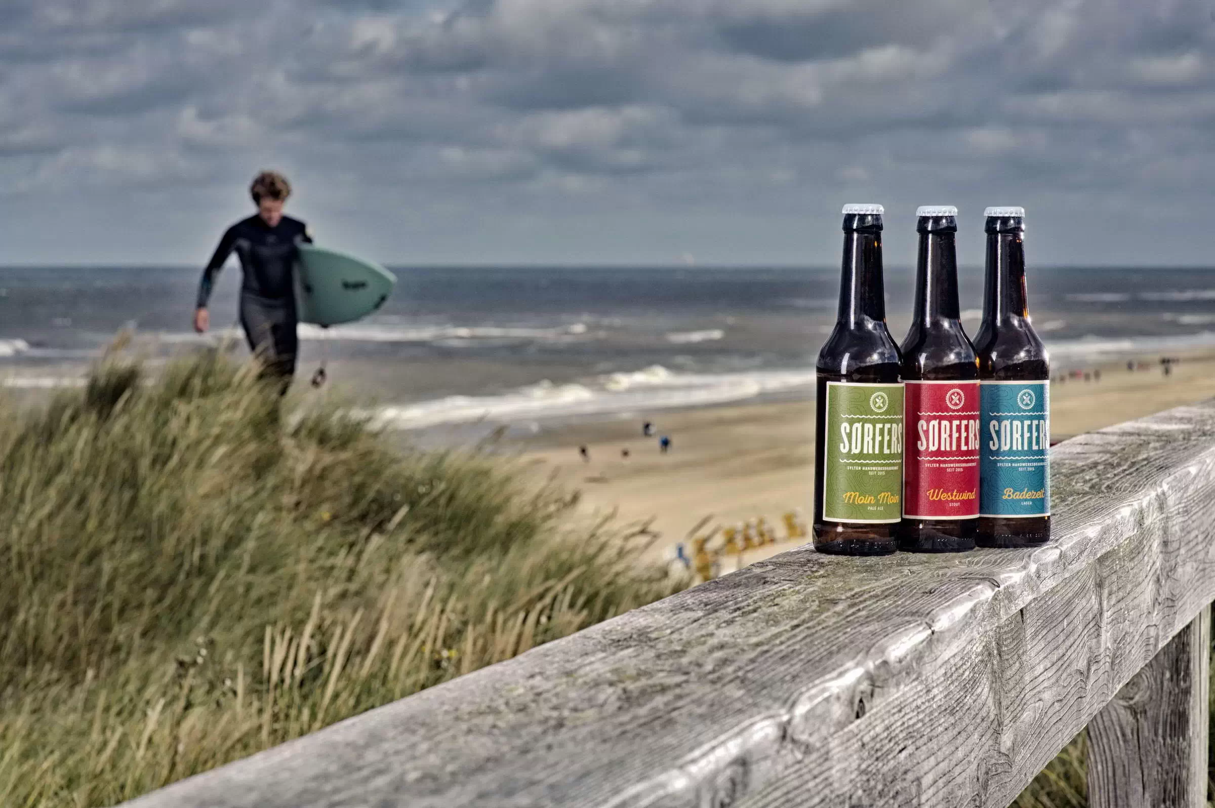 Drei SØRFERS Bierflaschen stehen auf einem Holzgeländer vor einer Sylter Dünenlandschaft mit einem Surfer im Hintergrund am Strand.