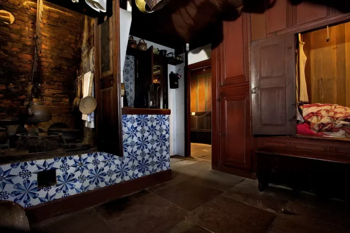 Historische Küche auf Sylt mit blau-weißen Kacheln, offenem Kamin und einer traditionellen Schrankbettnische in einem rustikalen Wohnraum.