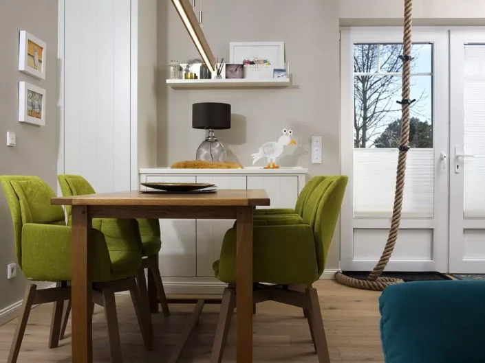 Ein gemütlicher Essbereich in einer Sylter Immobilie mit grünen Stühlen, Holztisch, maritimer Dekoration und einem dekorativen Tau an der Tür.