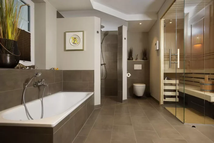 Modernes Badezimmer in einer Sylter Immobilie mit Badewanne, begehbarer Dusche, WC und einer integrierten Sauna mit Glasfront in eleganten Tönen.