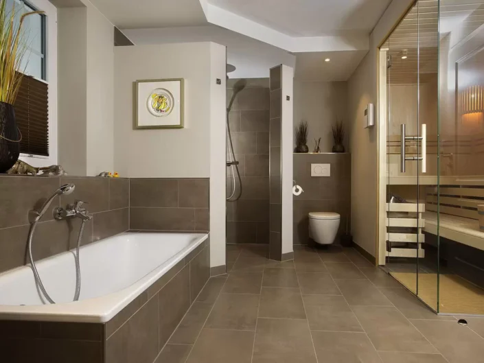 Modernes Badezimmer in einer Sylter Immobilie mit Badewanne, begehbarer Dusche, WC und einer integrierten Sauna mit Glasfront in eleganten Tönen.