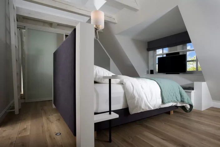 Modernes Schlafzimmer in einer Sylter Immobilie mit einem dunklen Bett, Holzboden und einem Fernseher unter einer Dachschräge mit Fenster.