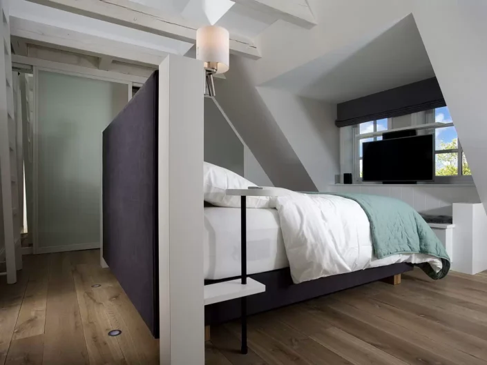 Modernes Schlafzimmer in einer Sylter Immobilie mit einem dunklen Bett, Holzboden und einem Fernseher unter einer Dachschräge mit Fenster.