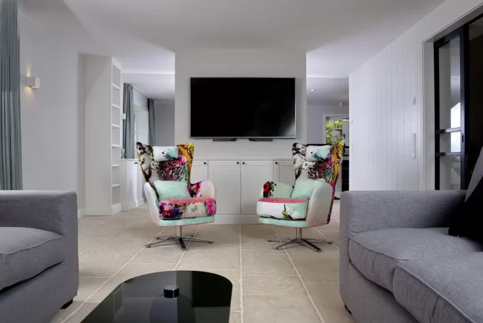 Modernes Wohnzimmer auf Sylt mit zwei bunten Sesseln, einem großen Flachbildfernseher und hellen Fliesenböden in einem eleganten Ambiente.