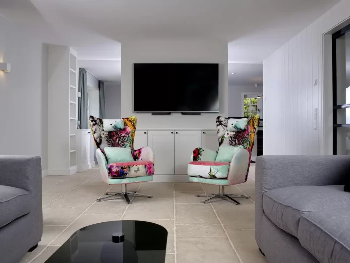 Modernes Wohnzimmer auf Sylt mit zwei bunten Sesseln, einem großen Flachbildfernseher und hellen Fliesenböden in einem eleganten Ambiente.