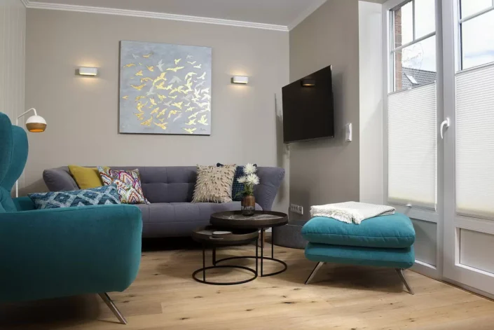 Modern eingerichtetes Wohnzimmer einer Sylter Immobilie mit grauem Sofa, türkisfarbenem Sessel, Couchtischen und einem Wandbild mit Vogelmotiven.