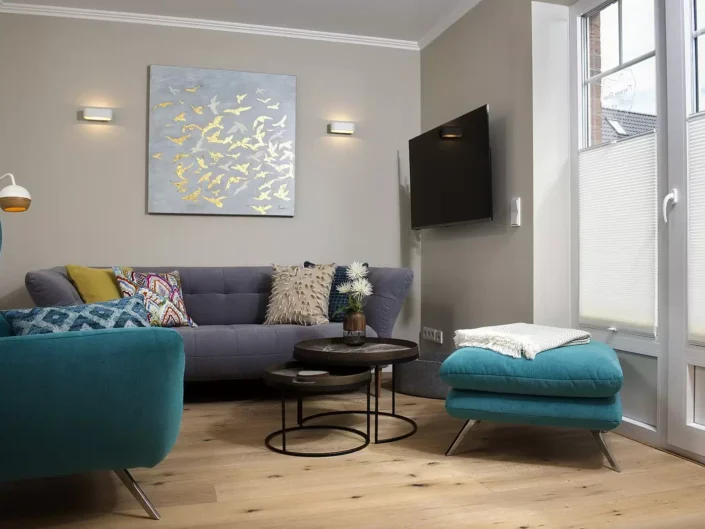 Modern eingerichtetes Wohnzimmer einer Sylter Immobilie mit grauem Sofa, türkisfarbenem Sessel, Couchtischen und einem Wandbild mit Vogelmotiven.