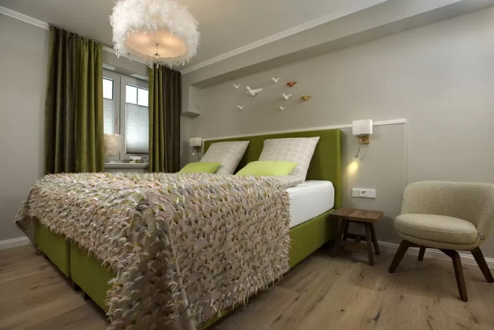 Modernes Schlafzimmer in einer Sylter Immobilie mit grünem Polsterbett, flauschiger Tagesdecke, Federlampe und dekorativen Vögeln an der Wand.